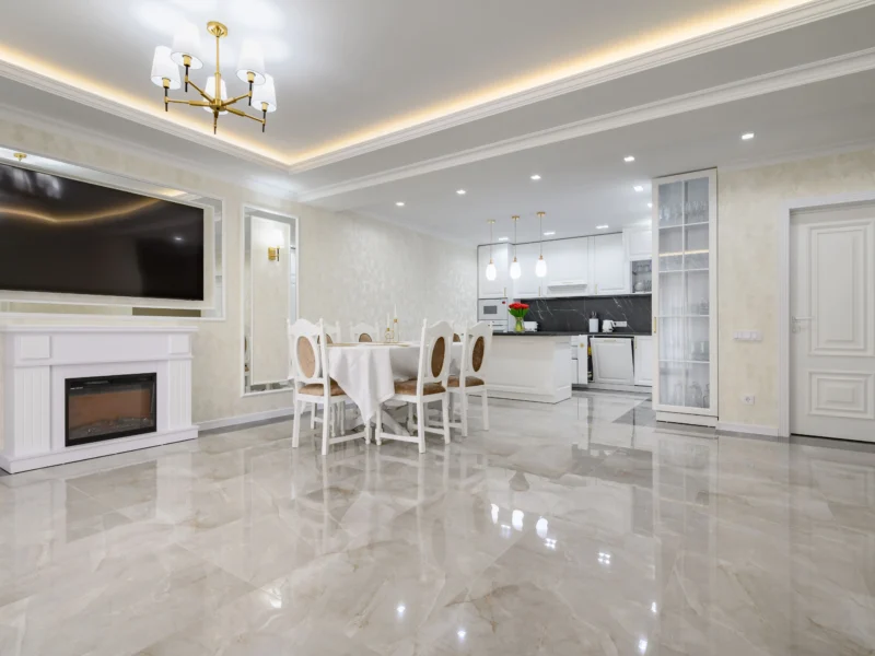 A spacious, luxurious open-plan living area featuring polished marble flooring, a white electric fireplace with a TV mounted above, a classic dining set, and a modern white kitchen in the background
